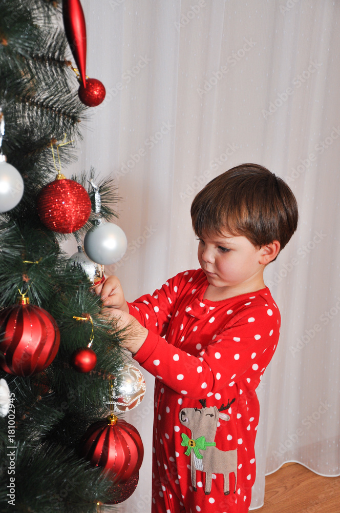 Boy in New Year's pajama decorating Christmas tree. Child putting decorations on Christmas tree Boy in New Year's pajama decorating Christmas tree. Child putting decorations on Christmas tree