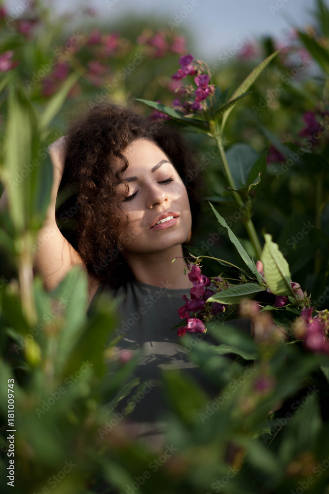 Curly brunette enjoys the rays of the sun among the pink flowers. Medicinal plant Impatiens glandulifera or Himalayan Balsam. Green leaves . Curly brunette enjoys the rays of the sun among the pink flowers. Medicinal plant Impatiens glandulifera or Himalayan Balsam. Green leaves .