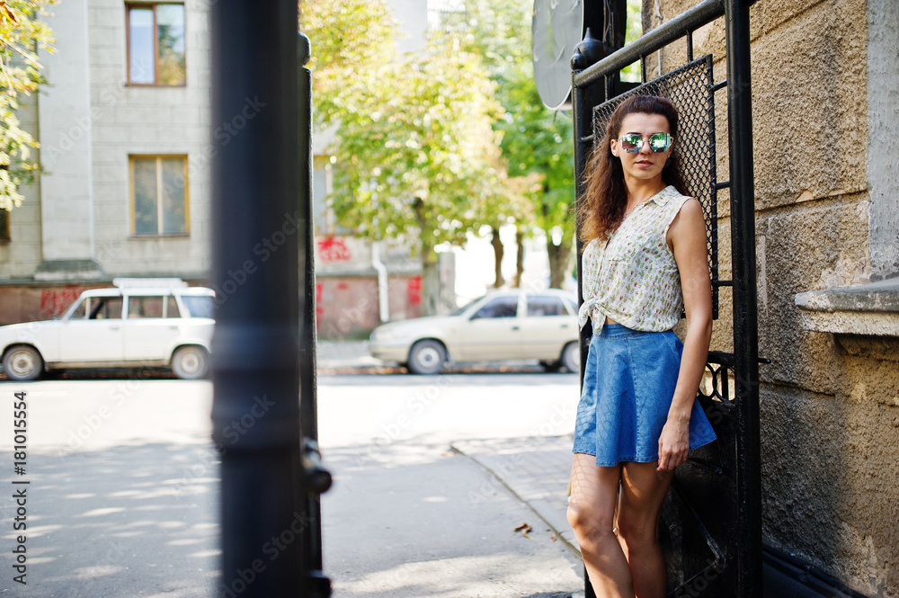 Curly stylish girl wear on blue jeans skirt, blouse and sunglasses. Portrait on streets of city.