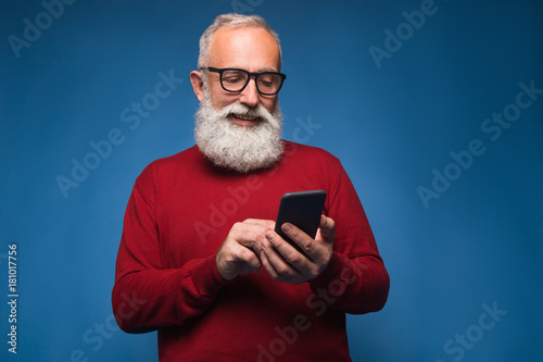 Using internet on smartphone isolated on blue background. Happy typing a new message to friends by bearded man in red sweeter and fashion glasses © stacestock