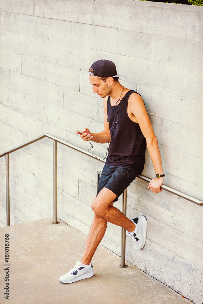 Man summer casual street fashion in New York. Wearing black tank top,  shorts, white sneakers, cap worn backward, a young guy stands against wall,  reading messages on cell phone. Filtered effect.. Stock