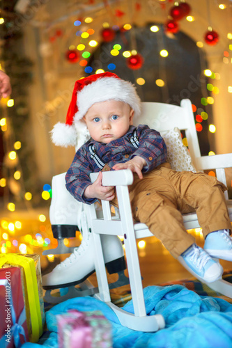a small funny kid in a Santa Claus hat sits on a chair, a set of bright festive lights in the background..