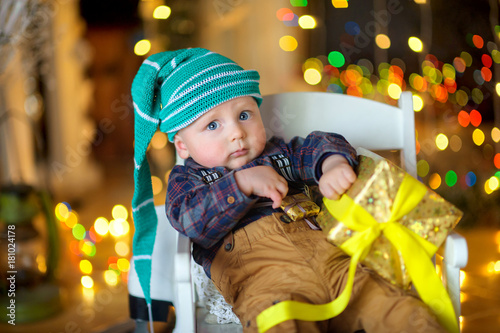 little funny baby in a hat dwarfs sitting on a chair with a gift in his hands, a set of bright festive lights in the background..