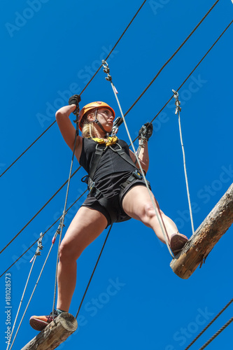 young girl in a mountain helmet walks at a height on wooden logs with ropes in an alpinist adventure park against a blue sky. training mountaineers in the mountains. rope park. leisure in nature