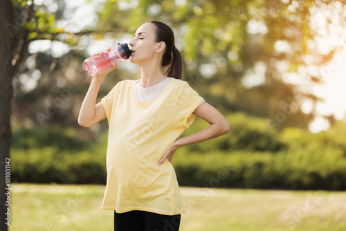 Pregnant woman walking in the park with a sports bottle in hands. She drinks from a bottle