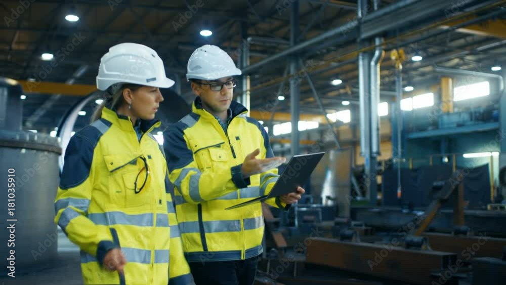 Male and Female Industrial Engineers in Hard Hats and Safety Jackets Discuss New Project while Using Laptop. 
