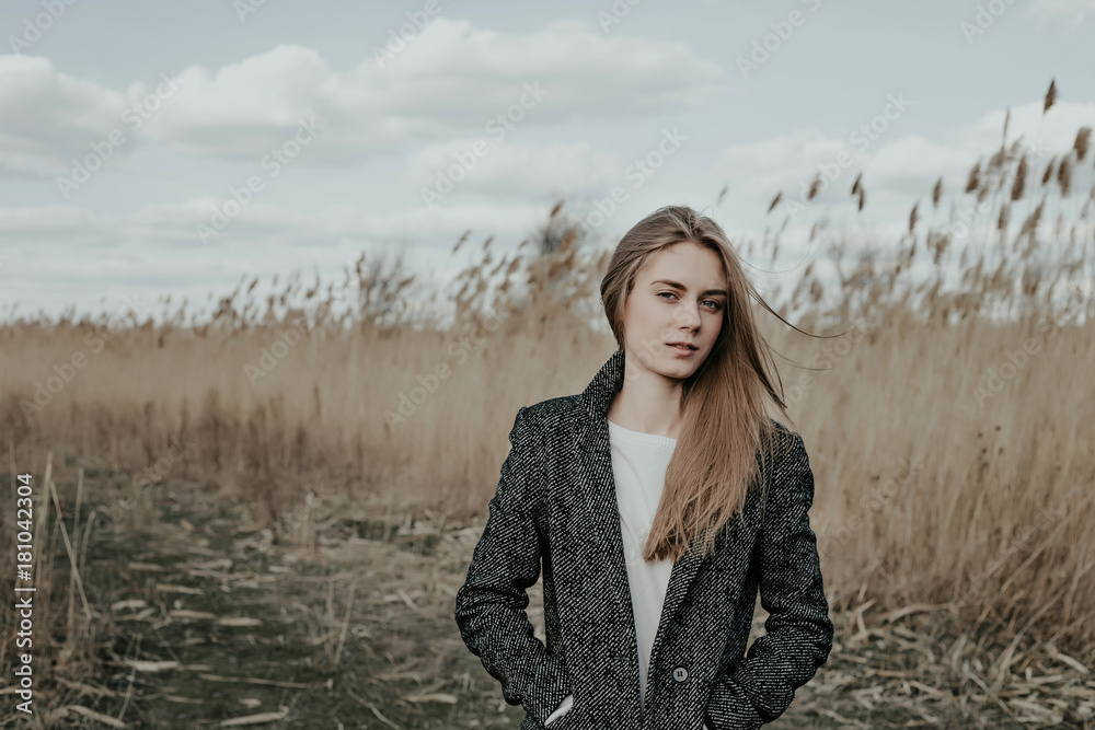 Beautiful woman dressed in coat and white sweater standing on bulrash background and looking at camera with screwing up one's eyes. Outdoor. Vouge. Copy space. Beautiful woman dressed in coat and white sweater standing on bulrash background and looking at camera with screwing up one's eyes. Outdoor. Vouge. Copy space.