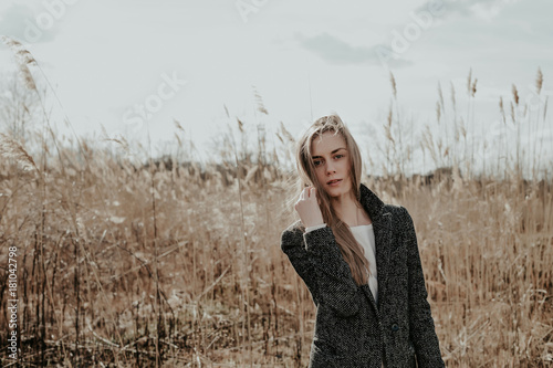 Woman looking at camera and touching to her hair. Girl with cute face, wide eyebrows and blonde hair posing outdoor. Background bulrush. Outdoor. Medium shot. Copy space.