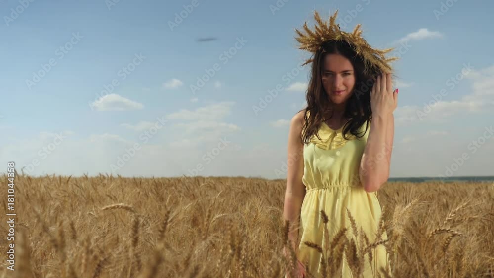 Woman in wheat crown and yellow dress looks up to camera. Field and blue sky Woman in wheat crown and yellow dress looks up to camera. Field and blue sky
