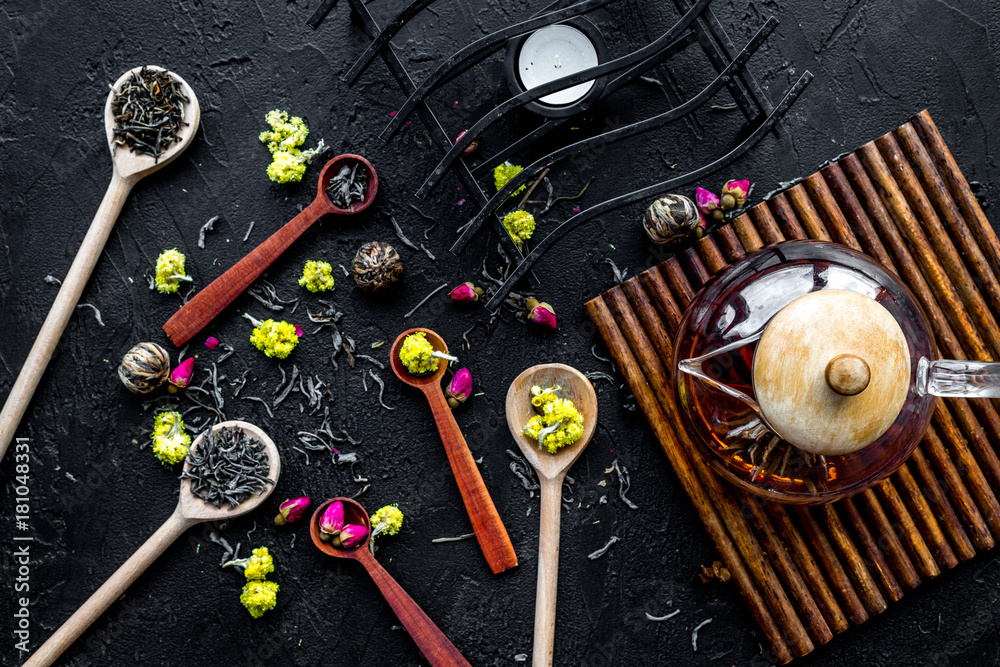 Brew the aromatic tea. Tea pot near wooden spoons with dried tea leaves, flowers and spices on black wooden background top view