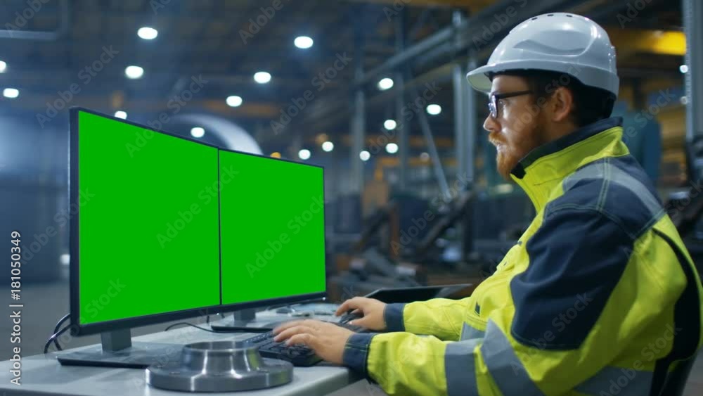 Inside the Heavy Industry Factory Industrial Engineer Works on the Personal Computer with Two Green Mock-up Screens. Inside the Heavy Industry Factory Industrial Engineer Works on the Personal Computer with Two Green Mock-up Screens.