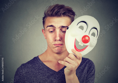 Young sad man taking off happy clown mask isolated on gray background. Human emotions. photo