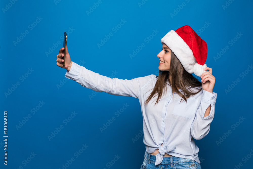 Obraz premium Santa girl making funny selfy on phone. Christmas hat isolated portrait of a woman on a blue background.