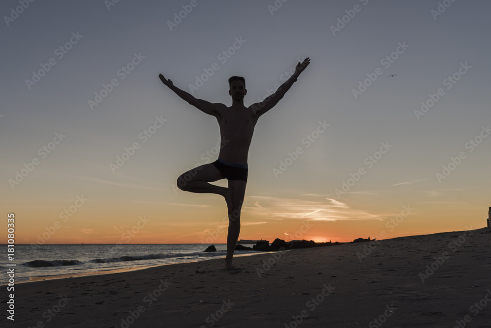 Silhouetted man doing tree yoga pose at sunset on the beach