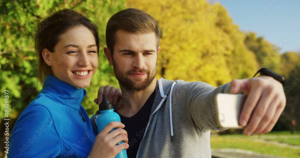 Close up of young smiling couple of runners taking selfies and showing victory signs in the sunny park in early autumn. Outside. Portrait shot Close up of young smiling couple of runners taking selfies and showing victory signs in the sunny park in early autumn. Outside. Portrait shot