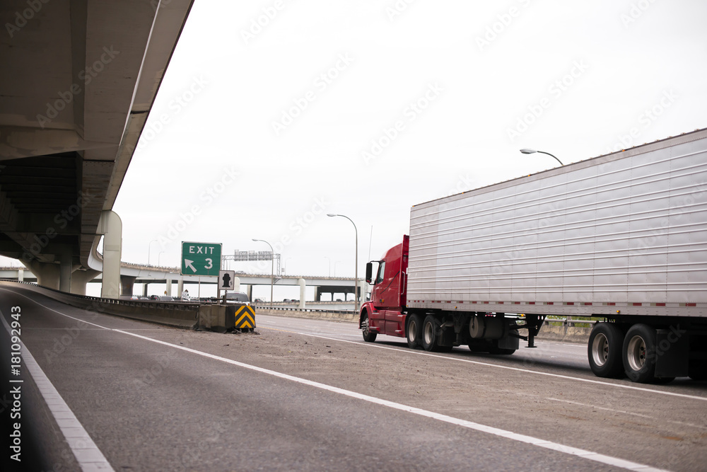 Red big rig semi truck with long reefer trailer transporting frozen cargo by interstate highway with overpass bridge