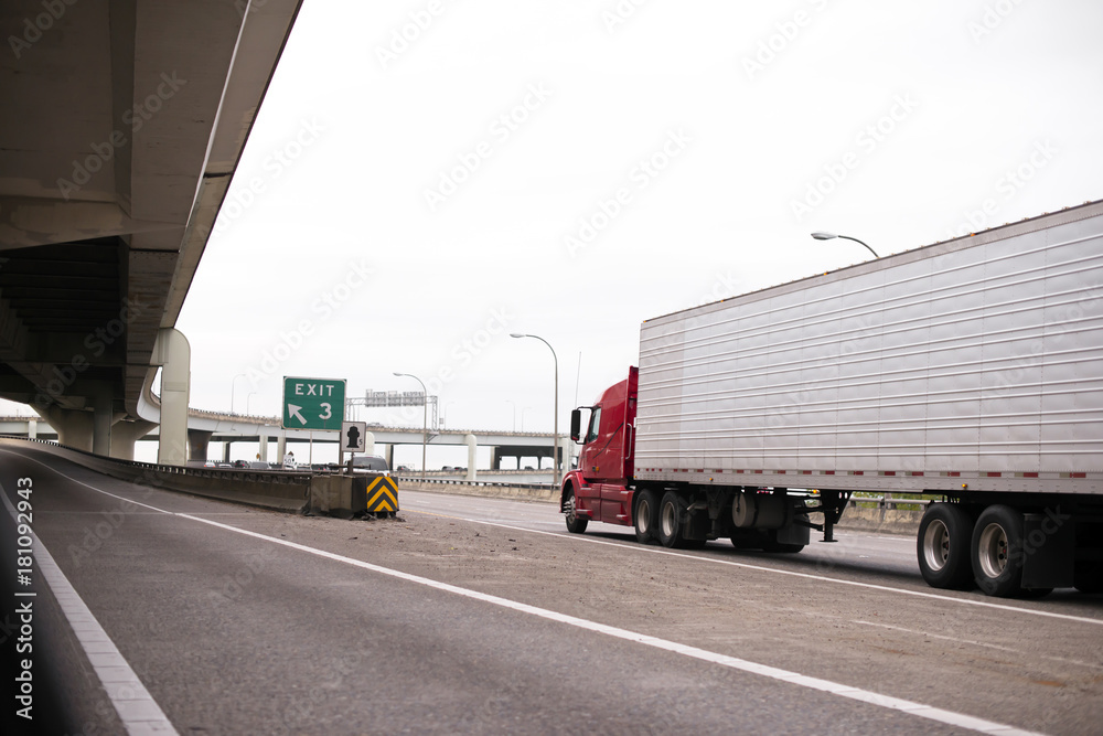 Red big rig semi truck with long reefer trailer transporting frozen cargo by interstate highway with overpass bridge Red big rig semi truck with long reefer trailer transporting frozen cargo by interstate highway with overpass bridge