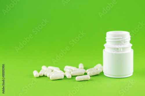 Close up white pill bottle with spilled out capsules on green background with copy space. Focus on foreground, soft bokeh. Pharmacy drugstore concept