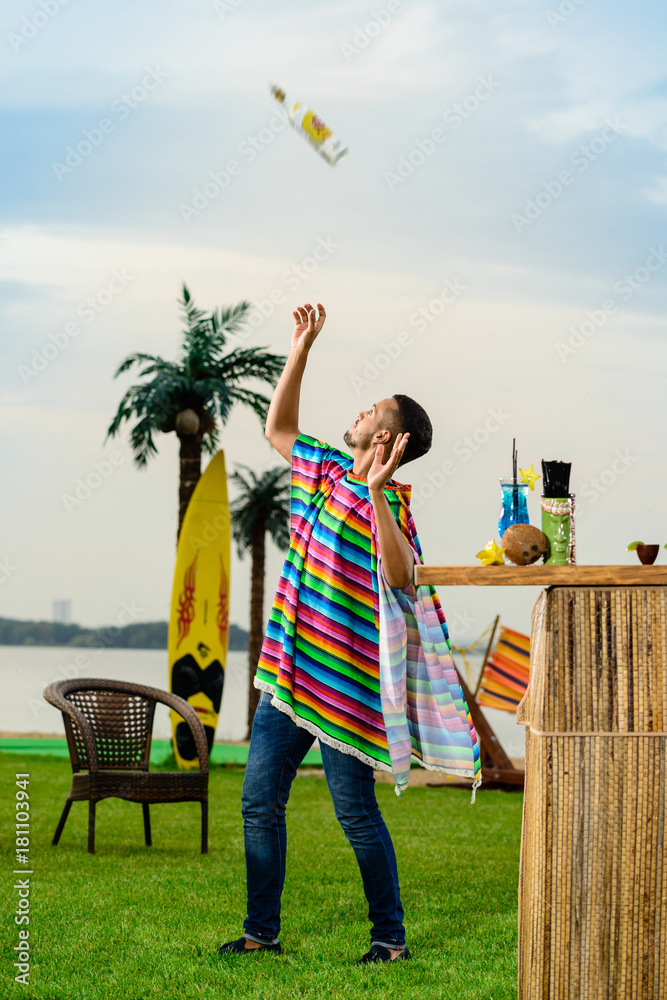 Handsome positive Mexican bartender in colorful poncho throws up a bottle  of alcohol near the bar counter at the resort Stock Photo | Adobe Stock