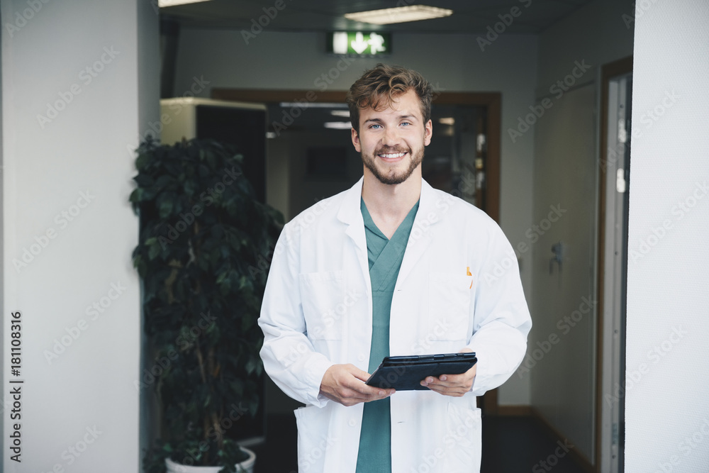Portrait of confident smiling male doctor standing with digital tablet at hospital corridor Portrait of confident smiling male doctor standing with digital tablet at hospital corridor