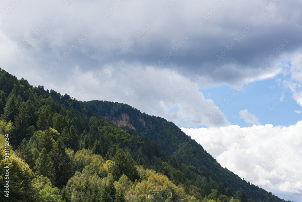 Mountain peaks covered with forests in various shades of green, in the background a very cloudy sky, Alps