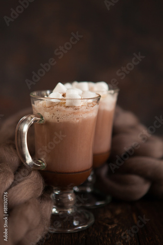 Two glasses with Hot chocolate garnished with whipped cream, marsmallow and cocoa powder on  wooden background with Cozy plaid from merinose wool