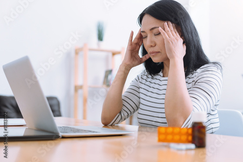 Trying to concentrate. Serious female person keeping eyes closed and putting elbows on the table while sitting opposite her laptop