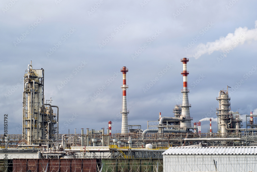general view of a chemical or oil refinery with a multitude of pipelines, factory pipes, distillation columns and freight cars in the foreground under a cloudy sky..