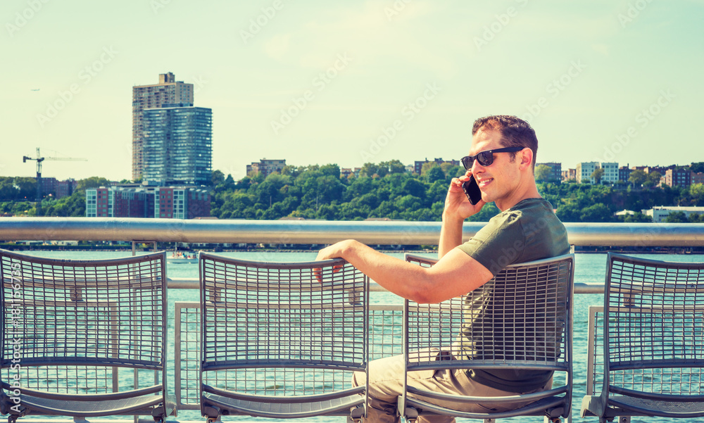 American Man summer vacation in New York. A young professional wearing green T shirt, sunglasses, sits on chair by Hudson River in New York, facing New Jersey, listening, talking on cell phone.. American Man summer vacation in New York. A young professional wearing green T shirt, sunglasses, sits on chair by Hudson River in New York, facing New Jersey, listening, talking on cell phone..