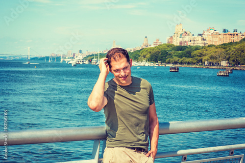 American Man summer vacation in New York. A young guy wearing green T shirt, stands by metal fence by Hudson River, looking down, scratching head, smiling, thinking, Bridge, harbor on background..