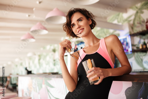 Young joyful girl in trendy swimsuit leaning on beach bar counter with sunglasses and cocktail in hands. Portrait of beautiful smiling lady standing at bar counter and dreamily looking aside