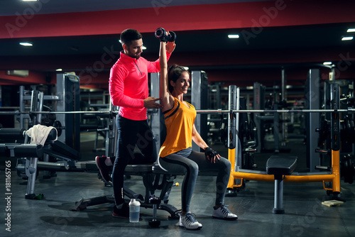 Pretty attractive fitness girl doing shoulder exercise with her personal trainer in the gym.