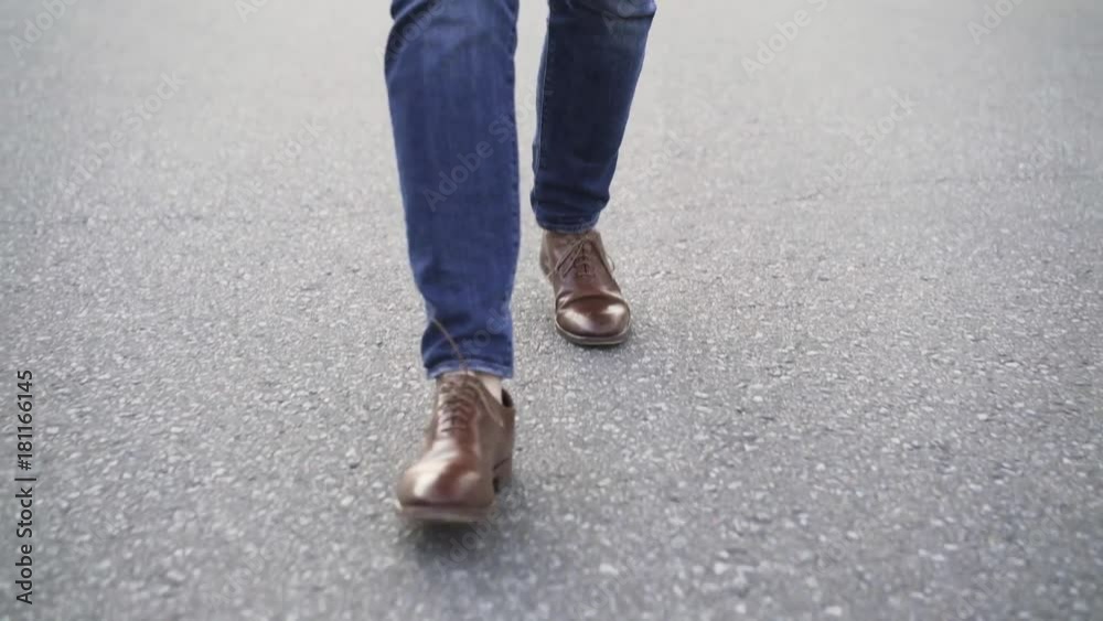 Unrecognizable man wearing blue jeans and brown leather shoes is walking in the street with no traffic. Tracking real time establishing shot Unrecognizable man wearing blue jeans and brown leather shoes is walking in the street with no traffic. Tracking real time establishing shot