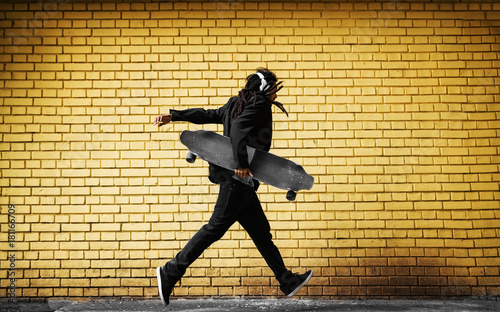 Handsome young dreadlocks skater with headphones running in a suit near the yellow wall on the street.