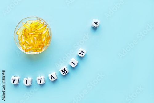Vászonkép Yellow capsules in the round glass bowl and the word vitamin A from white cubes with letters on a blue background