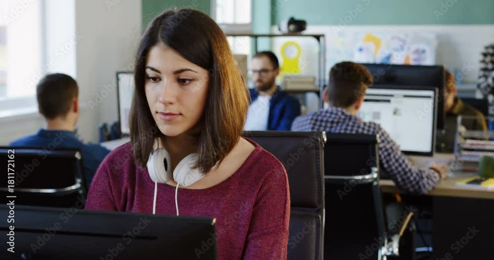 Portrait shot of young attractive woman with headphones working on the computer and smiling in the modern office space. Office workers on the background. Close up.