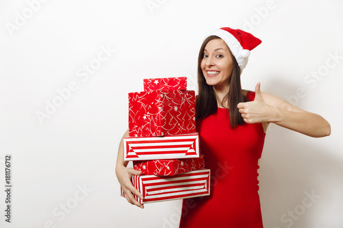 Smiling caucasian young happy woman dressed in red dress and Christmas hat holding gift boxes and showing thumb up on white background. Santa girl with present isolated. New Year holiday 2018 concept photo