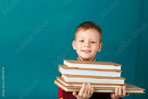 Cheerful thoughtful little school boy in school uniform with backpack and big pile of books standing against blue wall. Looking at camera. School concept.