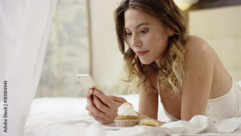 Woman lying on bed near cupcake and watching video on smartphone Woman lying on bed near cupcake and watching video on smartphone