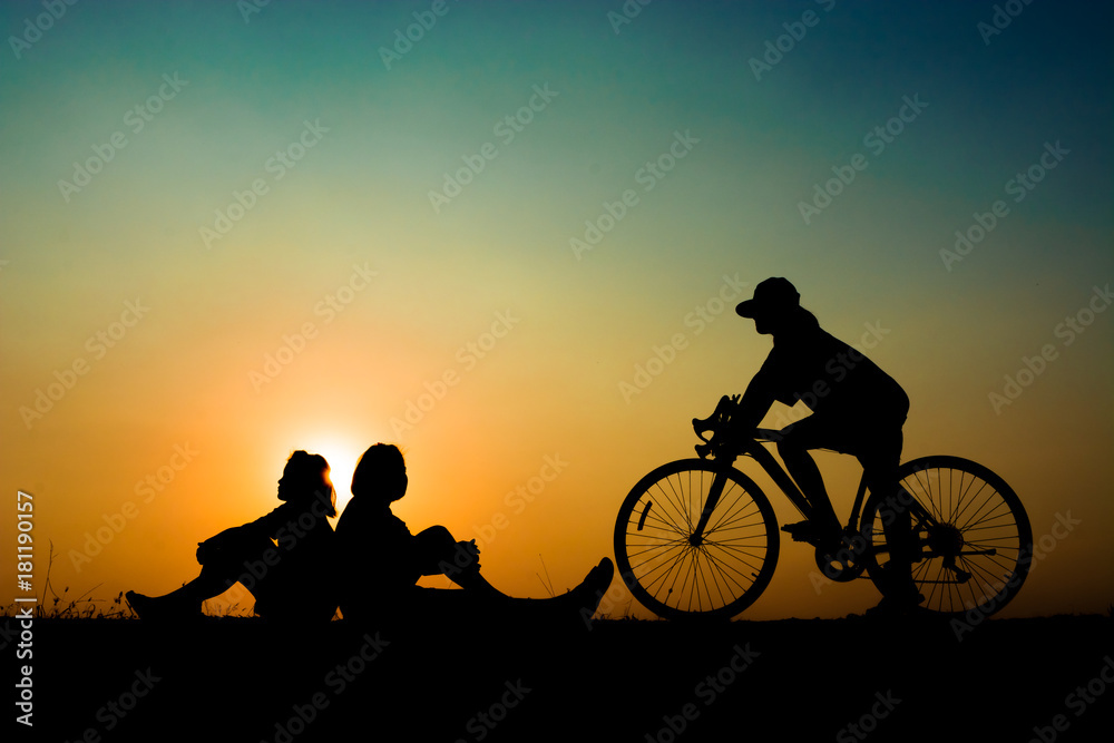 Obraz premium A silhouette of two people sitting on a hill with a person on a bicycle riding past them, all in front of a setting sun.