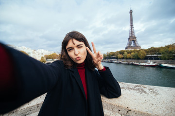 Beautiful young girl taking selfie with her phone making funny faces near the Eiffel tower in Paris