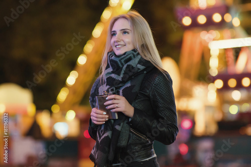 young beautiful woman alone drinks whiskey alcohol from flask. Outdoors at night in an amusement park. Night city background 