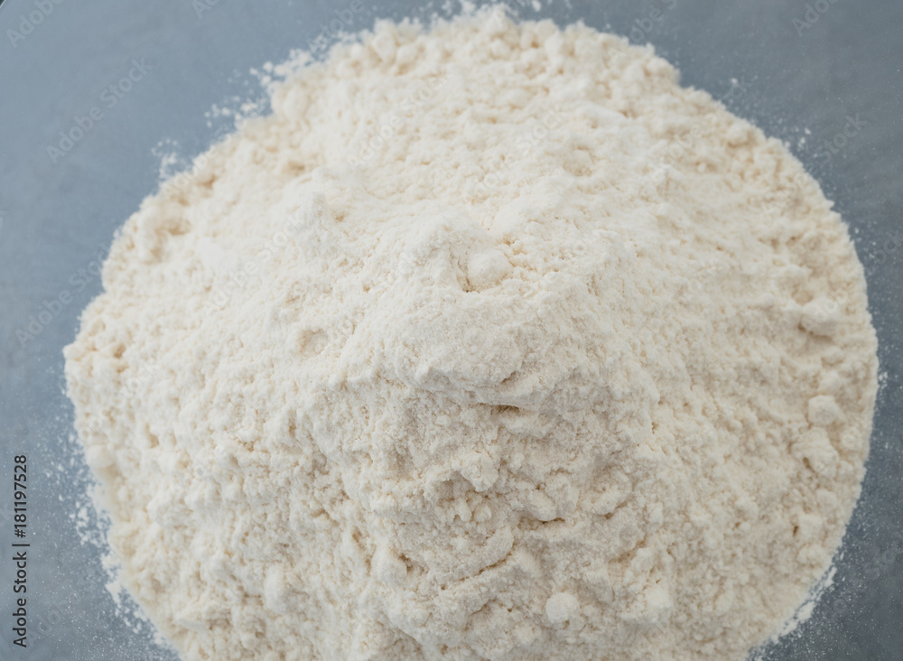 Close up shot of pile of all purpose white wheat flour in glass mixing bowl prepared for baking on blue background