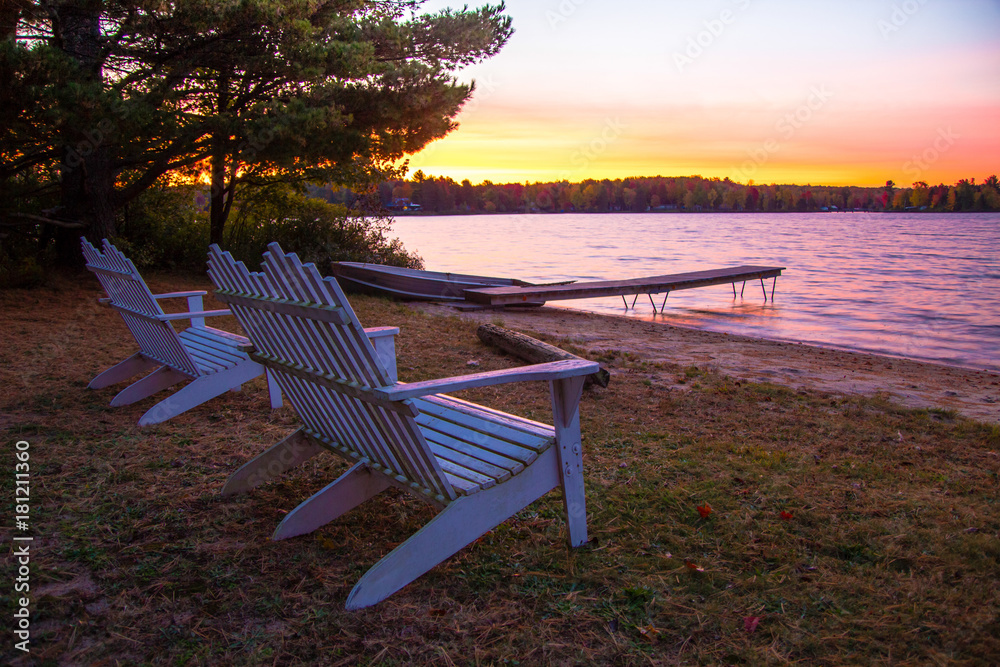 Summer Vacation At The Lake. Sunset over a lake with a row of Adirondack Chairs, dock and aluminum rowboat. - obrazy, fototapety, plakaty 