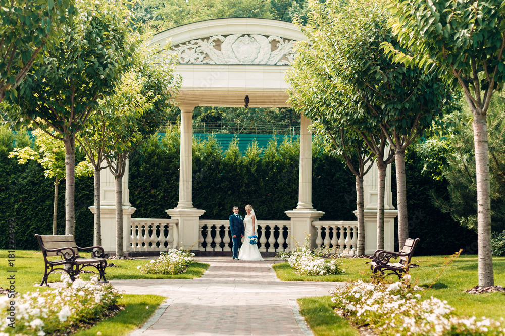 a beautiful young fair-haired bride stands next to the groom in an exotic park, in a long white dress with a bouquet of flowers in her hands, a walk after the wedding ceremony, a wedding ceremony