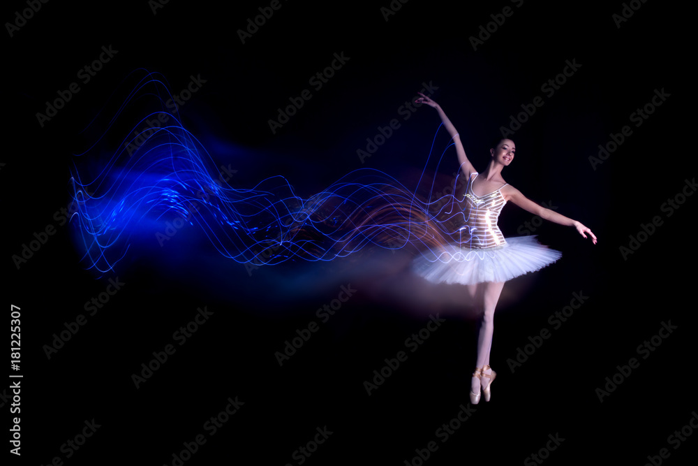 Young female / woman / girl ballerina in a white pack / tutu solo dancing doing stand on toes and leaves blue light leak trail of silhouette in black scene with reflecting floor and dark background