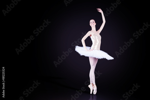 Young female / woman / girl ballerina in a white pack / tutu solo dancing and doing stand on toes in dark black scene with reflecting floor and dark background
