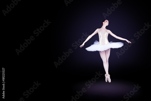 Young female / woman / girl ballerina in a white pack / tutu solo dancing and doing jump in dark black scene with reflecting floor and dark background