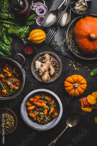 Vegan pumpkin stew dish with spinach served in bowl with spoon on dark kitchen table background with pot and ingredients, top view, vertical. Healthy seasonal food and clean eating concept