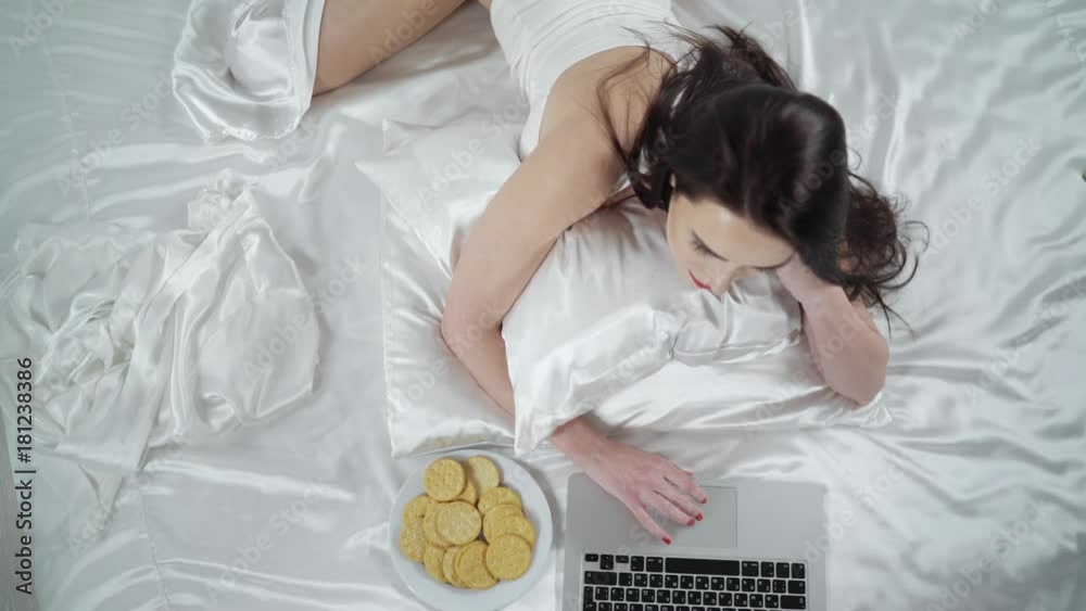 Beautiful woman in bedroom lying on the bed working with laptop. Young brunette woman in underwear lies across the white bed sheets on her stomach typing on notebook keyboard. View from above.